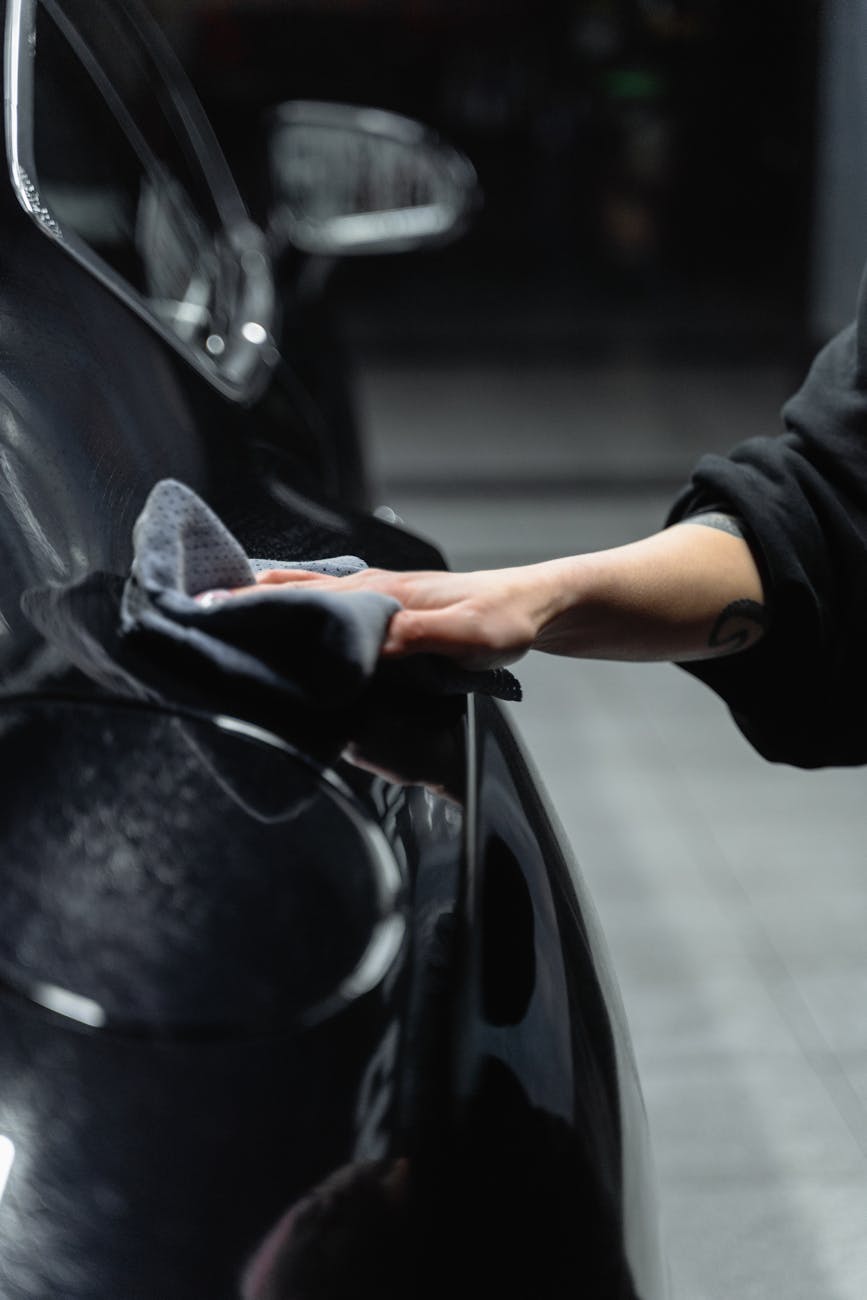 Close-up of a person's hand cleaning a shiny black car with a microfiber cloth in a garage.
